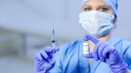 A doctor in medical gloves and a face mask in blur holds a vial with a vaccine and a syringe in his hands.