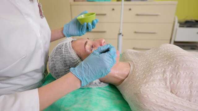 Professional beautician in gloves doing chemical peel for young woman, applying fruit acid with brush from bowl on face skin of female client during professional peeling procedure in beauty clinic