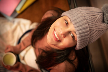 Top view of a happy young woman enjoying coffee in winter and looking at camera with a toothy smile.