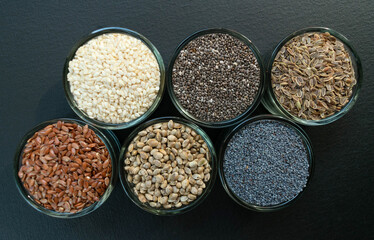 An assortment of healthy seeds (chia, hemp flax, dill, sesame, poppy seeds) - top view of small glass bowls against a black background.