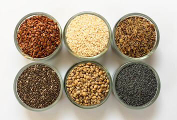 An assortment of healthy seeds (chia, hemp flax, dill, sesame, poppy seeds) - top view of small glass bowls on a white background.