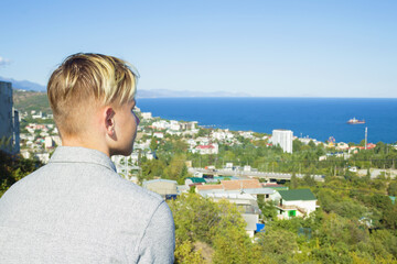 Fototapeta na wymiar A lone blond teenager in a gray shirt looks down at the coastal town of Alushta on the Crimean peninsula. The city is buried in greenery. Blue, cloudless sky and blue Black Sea. Mountains are visible.