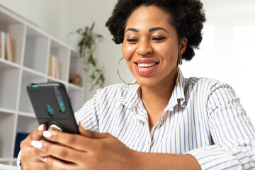 Portrait of a happy African American woman using mobile phone at home. Black smiling female chatting holding a smartphone in her hand in a light interior.