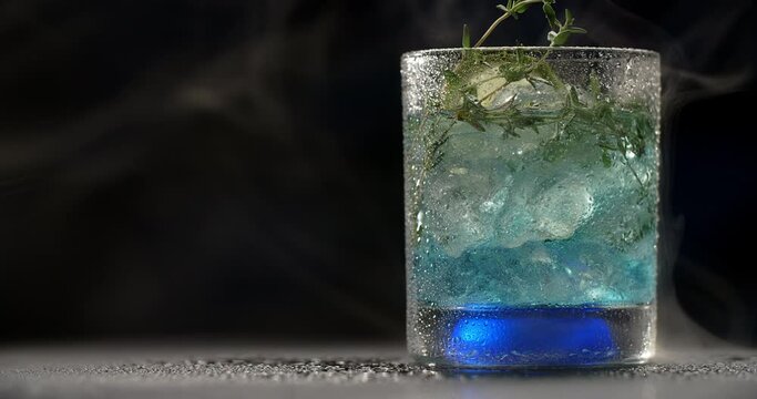 Cold glass with blue cocktail is spinning slowly in the smoke on the dark background, alcohol drinks and beverage, non-alcohol cocktail, object video of lemonade, 4k Prores HQ