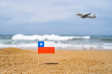 Flag of Chile in the form of a toothpick in the sand of beach opposite sea wave with landing airplane. Travel concept