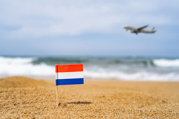 Flag of Netherlands in the form of a toothpick in the sand of beach opposite sea wave with landing airplane. Travel concept