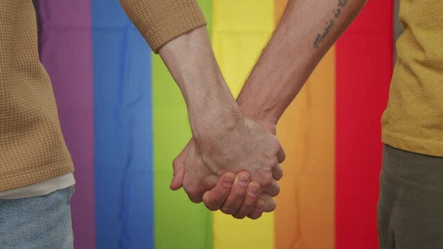 Close up shot of unrecognizable gay couple holding hands before rainbow flag