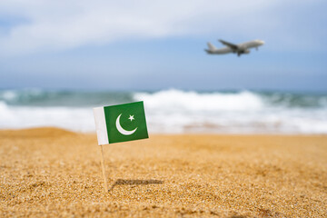 Flag of Pakistan in the form of a toothpick in the sand of beach opposite sea wave with landing airplane. Travel concept