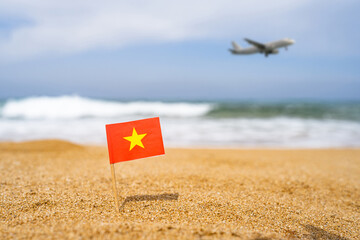 Fototapeta na wymiar Flag of Vietnam in the form of a toothpick in the sand of beach opposite sea wave with landing airplane. Travel concept