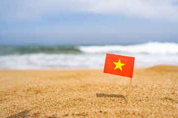Fototapeta na wymiar Flag of Vietnam in the form of a toothpick in the sand of beach opposite sea wave . Travel concept