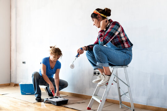 Two women renovating a house. Loading roller in paint tray. Sitting on painter ladder. Painting house walls white.
