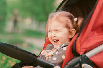 Screaming little toddler girl sitting in red buggy above defocused nature background and looking away.