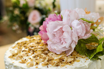 Delicious white cream cake, garnished with roasted almonds and edible pink peonies with green leaves. Banquet, celebration, confectionery. Close-up. Bokeh.