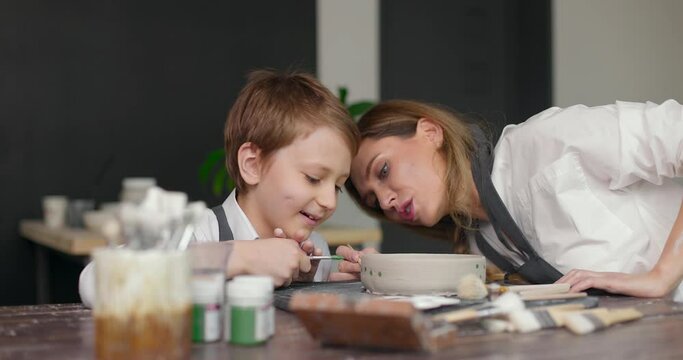 Pottery class workshop. Mother teaches her happy son drawing on clay bowl in pottery studio. Spending time together. Hobby, leisure. Slow motion