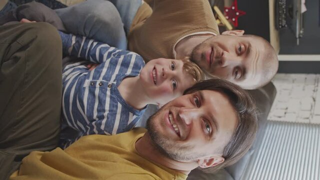 Vertical POV shot of happy gay fathers and little boy sitting together on couch at home and taking selfie