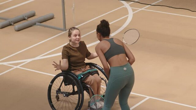High angle shot of young Mixed-Race woman and handicapped school-aged girl in wheelchair high-fiving and talking after winning match in indoor court