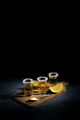 Dark photography of tequila shots with salt and lemon slices on a wooden board with copyspace.