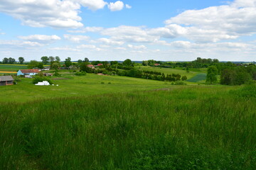 A view of a vast meadow, pastureland, or field covered with grass and some crops with some puffy clouds seen on a sunny summer day and some small shacks and village houses nearby in Poland