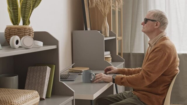 Side-view medium slowmo of blind senior man in black eyeglasses sitting by desk at home groping for cup of tea