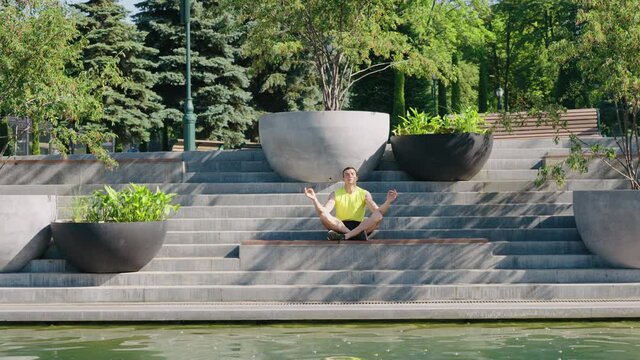 Man in sportswear sitting in lotus pose with closed eyes on staircase near pond. Tranquil yogi experiencing zen in rays of sunshine. Outdoor activity in summer. Concept of meditation