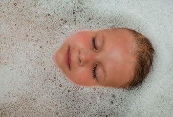 Little girl bathes in a bathtub, face in foam