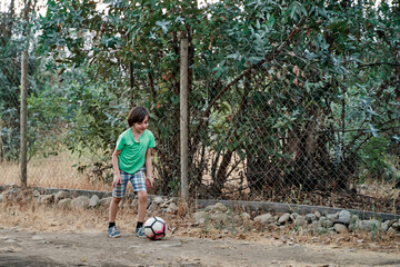 portrait of a little boy hitting a soccer ball in a field between trees and a dirt road, in the...