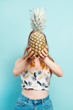 Cheerful young blonde holding a pineapple in front of her face with a summer top on a blue background. Summer concept. Vertical image with copy space