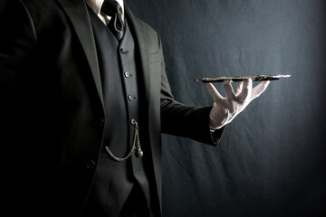 Portrait of Butler in Dark Suit and White Gloves Beautifully Holding Silver Serving Tray on Black Background. Concept of Service Industry and Professional Hospitality. 