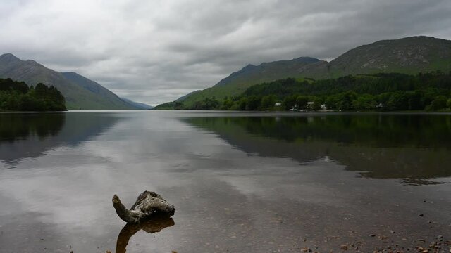 4k time lapse of cloud movement over Loch Shiel, Glenfinnan, Scottish Highlands on an overcast day. Cloud movement is reflected in calm water. Log in foreground.