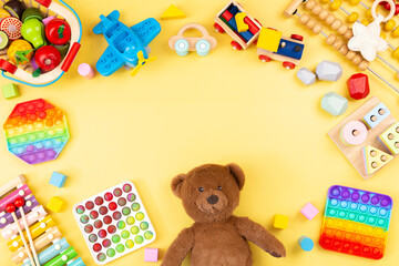 Baby kids toy frame with teddy bear, wooden and musical toys, abacus, plane, pop it fidget toys and colorful blocks on yellow background. Top view, flat lay - obrazy, fototapety, plakaty