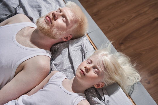 Girl laying at the bed with closed eyes near her bearded albino father and relaxing