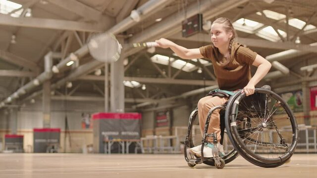 Long shot of blond-haired Caucasian handicapped sportsgirl in wheelchair playing badminton, serving shuttlecock with racket in indoor court