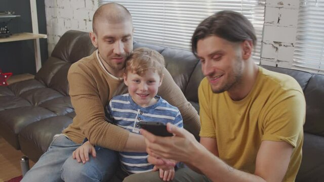 Handheld slowmo of cheerful gay fathers and their cute little son sitting together on couch and taking selfie on mobile phone