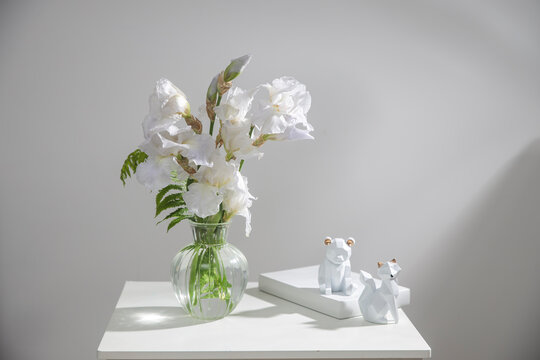 A bouquet of three white irises and a fern in a transparent vase on the table. Two ceramic figurines of a fox and a bear