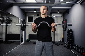 Fototapeta na wymiar Hand training with sports equipment. A portrait of half the body of a handsome and muscular man in the gym exercising his biceps with a barbell. Modern gym with a dark atmosphere, sports life