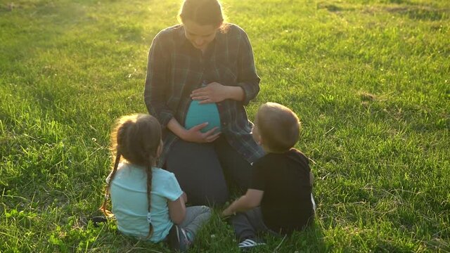 summer, nature, happy family, pregnancy - young pregnant woman mother with two small toddler children sit on grass in park at sunset. Kids stroking mom belly kiss have fun, laugh spend time together