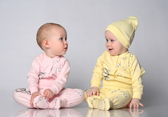 Two baby toddler babies are sitting next to each other on a light studio background. One toddler is dressed in a yellow cotton jumpsuit with a cute hat, the other in a pink sandbag. Children's clothes