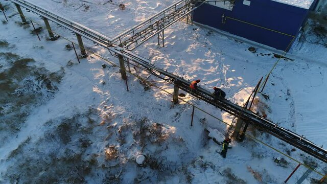 Workers repair outer pipeline near blue and white workshop building in snowy yard at sunset in winter evening aerial view