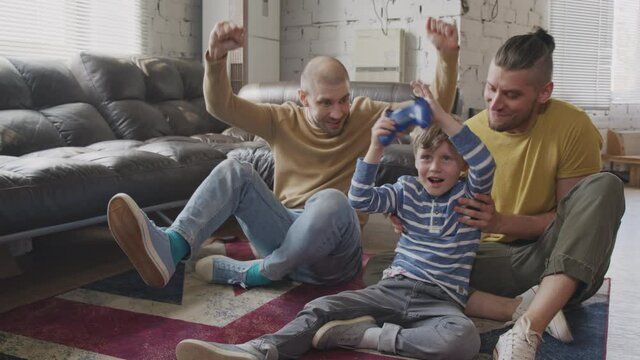 Slowmo shot of gay fathers sitting on rug on floor and watching their little son playing video game They are celebrating him winning