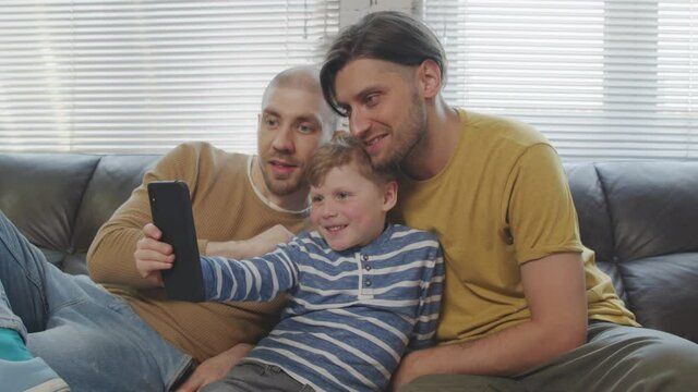 Slowmo shot of happy gay fathers and cute little son sitting on couch together and taking photo on mobile phone