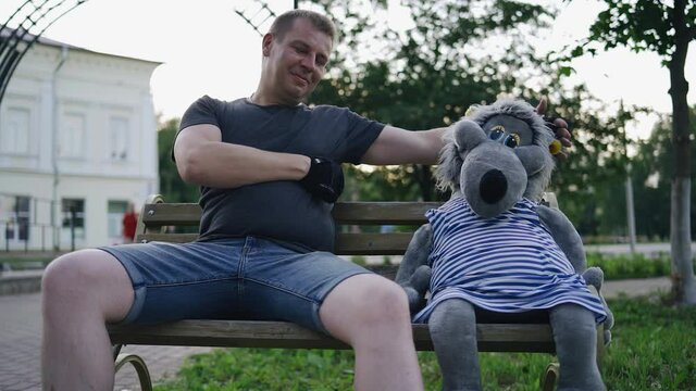 A man sits on a bench next to a stuffed wolf toy and puts his arm around it. Good mood