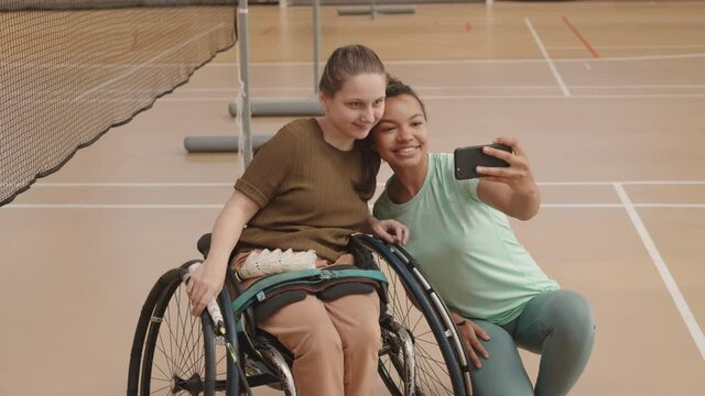 High angle medium long of young Mixed-Race woman and teenage Caucasian handicapped girl taking selfie with smartphone in indoor court
