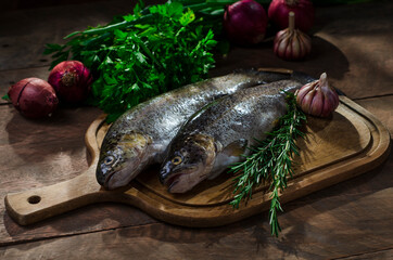 two trout on a wooden kitchen board on a rustic table with the sun's rays coming in through a window, some onions, garlic buboes and a sprig of rosemary