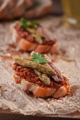 closeup of a bruschetta with dried tomato pesto, asparagus, sardines on a piece of brown paper with an unfocused background of another bruschetta, some pieces of Italian bread and olive oil