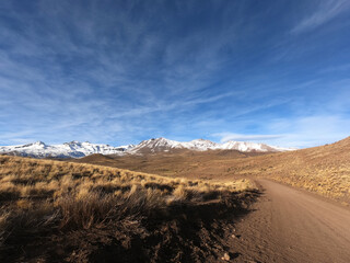 The dirt road across the golden valley and Andes cordillera. View of the rural route across the yellow grassland and hills, into Volcano Domuyo in Neuquén, Patagonia Argentina. 