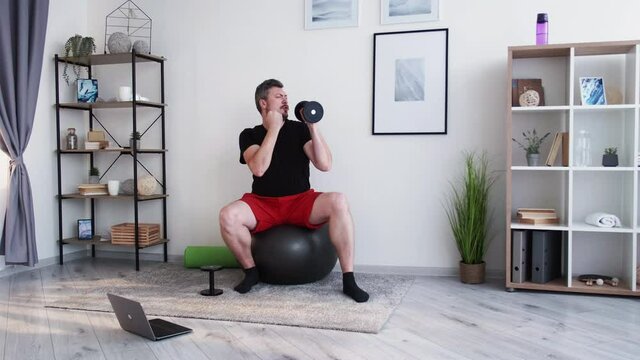Fitness performance. Funny fatty man. Online training. Home gym. Creative chubby guy sitting fit ball using dumbbells like microphone singing doing exercise on laptop in light room interior.