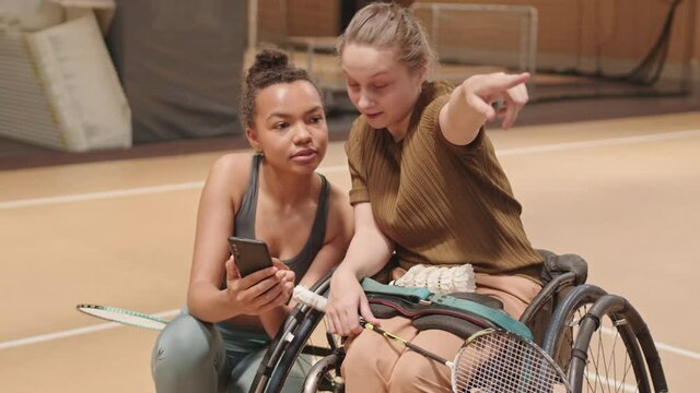 Medium long of teenage Caucasian girl in wheelchair talking to Mixed-Race female athlete showing pictures on smartphone, friends holding badminton rackets in gym
