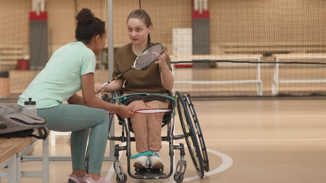 Locked-down of handicapped Caucasian girl on sports wheelchair talking to female partner by bench in gym, then both going away with badminton racket