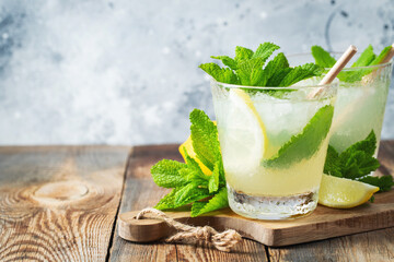 Two glass with lemonade or mojito cocktail with lemon and mint, cold refreshing drink or beverage with ice on rustic blue background. Copy space