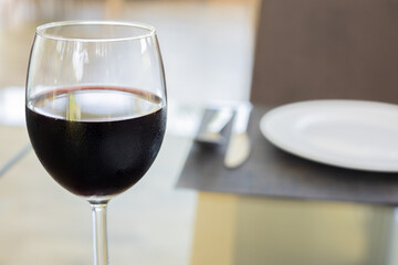 Selective focus and blurred background with copy space of glass of red wine with tableware clean set (white plate, fork and spoon) with prepared for food of lunch meal in luxury formal restaurant.